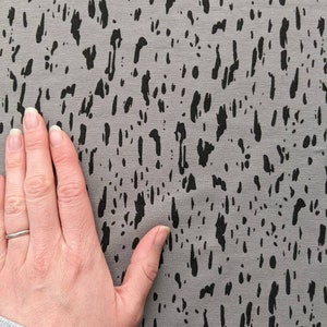K&ouml;nnte beinhalten: Nahaufnahme eines grauen Stoffes mit einem schwarzen abstrakten Druck. Der Druck &auml;hnelt kleinen, unregelm&auml;&szlig;igen Formen und Linien.