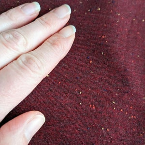 K&ouml;nnte beinhalten: Nahaufnahme einer Hand, die auf einem burgunderfarbenen Stoff mit einem gesprenkelten Muster aus kleinen roten, gelben und blauen Punkten liegt.
