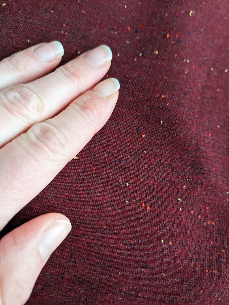 K&ouml;nnte beinhalten: Nahaufnahme einer Hand, die auf einem burgunderfarbenen Stoff mit einem gesprenkelten Muster aus kleinen roten, gelben und blauen Punkten liegt.