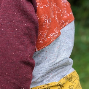 K&ouml;nnte beinhalten: Ein lang&auml;rmliges Shirt mit einem Patchwork-Design in Burgunderrot, Grau und Gelb. Der obere Teil des Shirts ist burgunderrot mit einem gesprenkelten Muster. Der mittlere Teil ist grau mit einem gesprenkelten Muster. Der untere Teil ist gelb mit einem wei&szlig;en Tierdruck.
