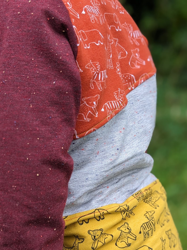 K&ouml;nnte beinhalten: Ein lang&auml;rmliges Shirt mit einem Patchwork-Design in Burgunderrot, Grau und Gelb. Der obere Teil des Shirts ist burgunderrot mit einem gesprenkelten Muster. Der mittlere Teil ist grau mit einem gesprenkelten Muster. Der untere Teil ist gelb mit einem wei&szlig;en Tierdruck.