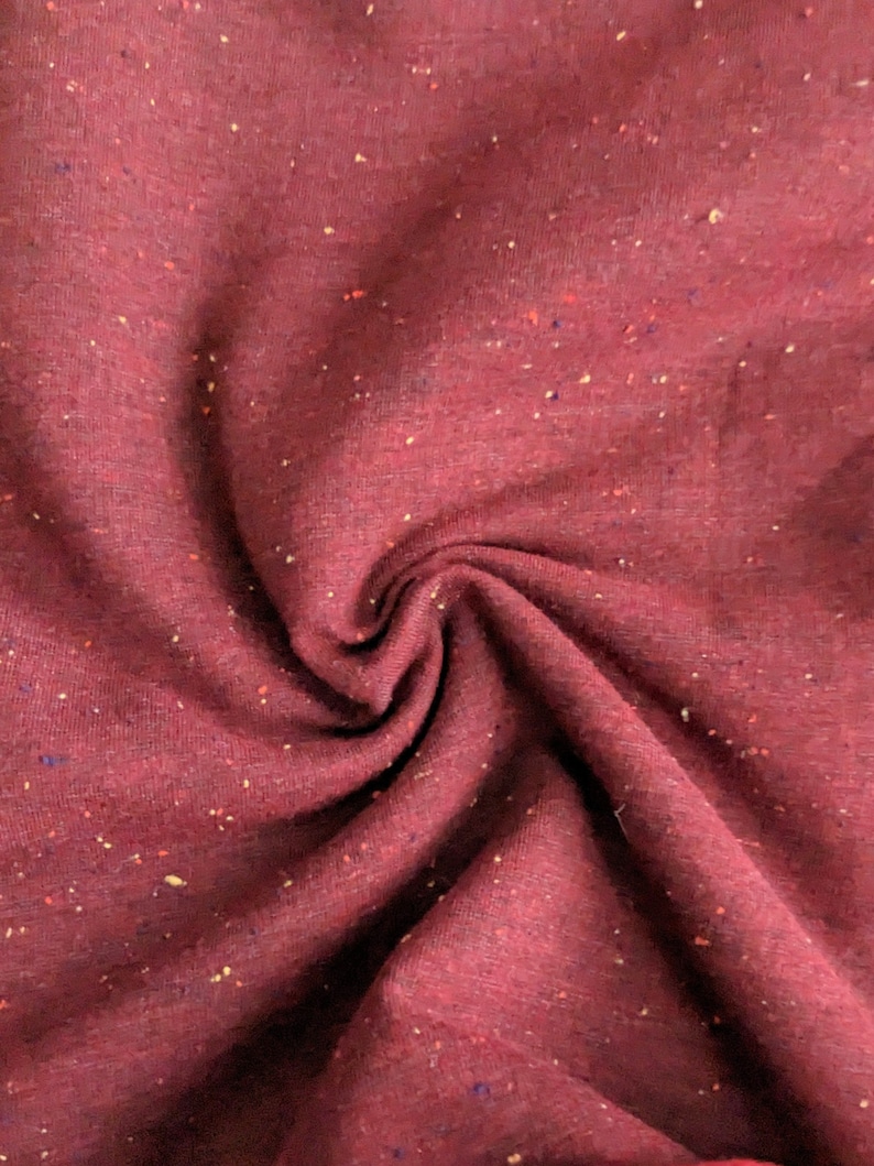 K&ouml;nnte beinhalten: Nahaufnahme eines burgunderfarbenen Stoffes mit einem gesprenkelten Muster aus kleinen gelben, roten und blauen Punkten. Der Stoff ist weich und texturiert.