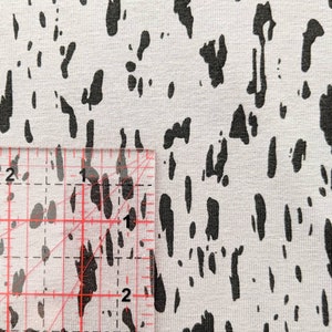 K&ouml;nnte beinhalten: Nahaufnahme eines wei&szlig;en Stoffes mit einem schwarzen abstrakten Druck. Ein Lineal liegt auf dem Stoff, um die Gr&ouml;&szlig;e des Drucks zu zeigen. Der Druck ist etwa 1,2 cm breit.
