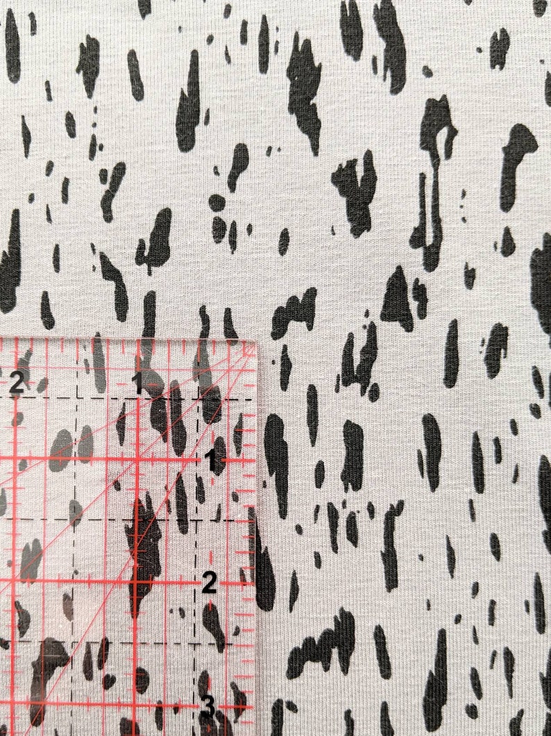 K&ouml;nnte beinhalten: Nahaufnahme eines wei&szlig;en Stoffes mit einem schwarzen abstrakten Druck. Ein Lineal liegt auf dem Stoff, um die Gr&ouml;&szlig;e des Drucks zu zeigen. Der Druck ist etwa 1,2 cm breit.