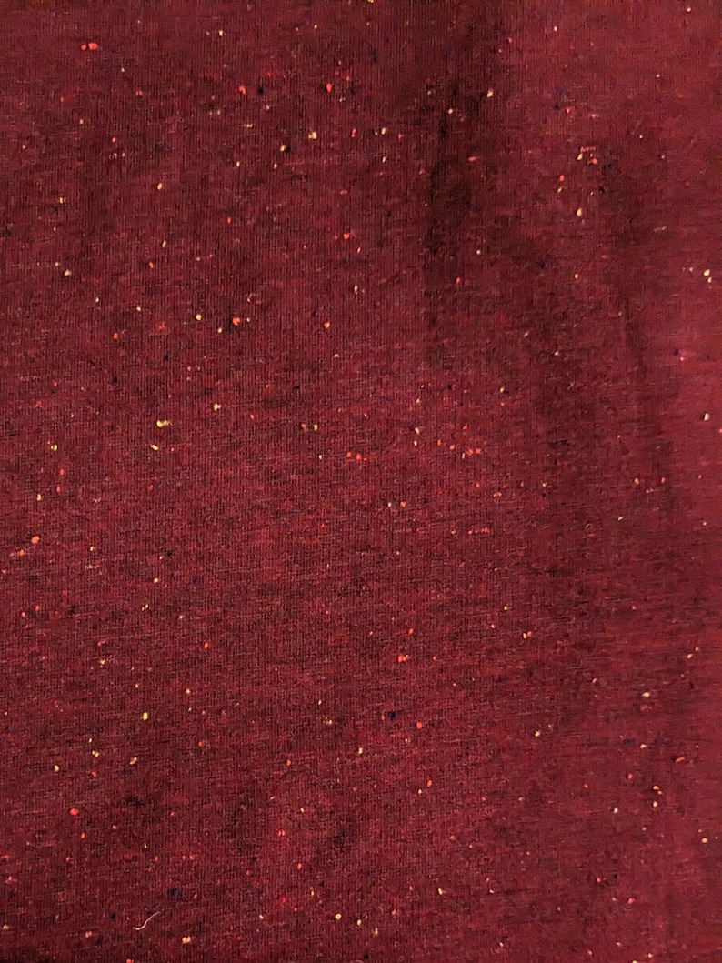 K&ouml;nnte beinhalten: Nahaufnahme eines tief roten Stoffes mit gesprenkelter Textur. Der Stoff hat einen dezenten Glanz und ist mit kleinen Flocken in Gelb, Orange und Blau gewebt.