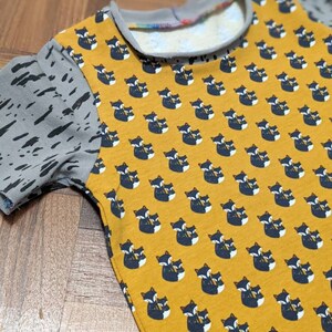 K&ouml;nnte beinhalten: Ein gelbes T-Shirt mit kurzen &Auml;rmeln und einem Muster aus schwarzen und wei&szlig;en F&uuml;chsen. Die &Auml;rmel sind aus grauem Stoff mit einem schwarzen Spritzmuster.