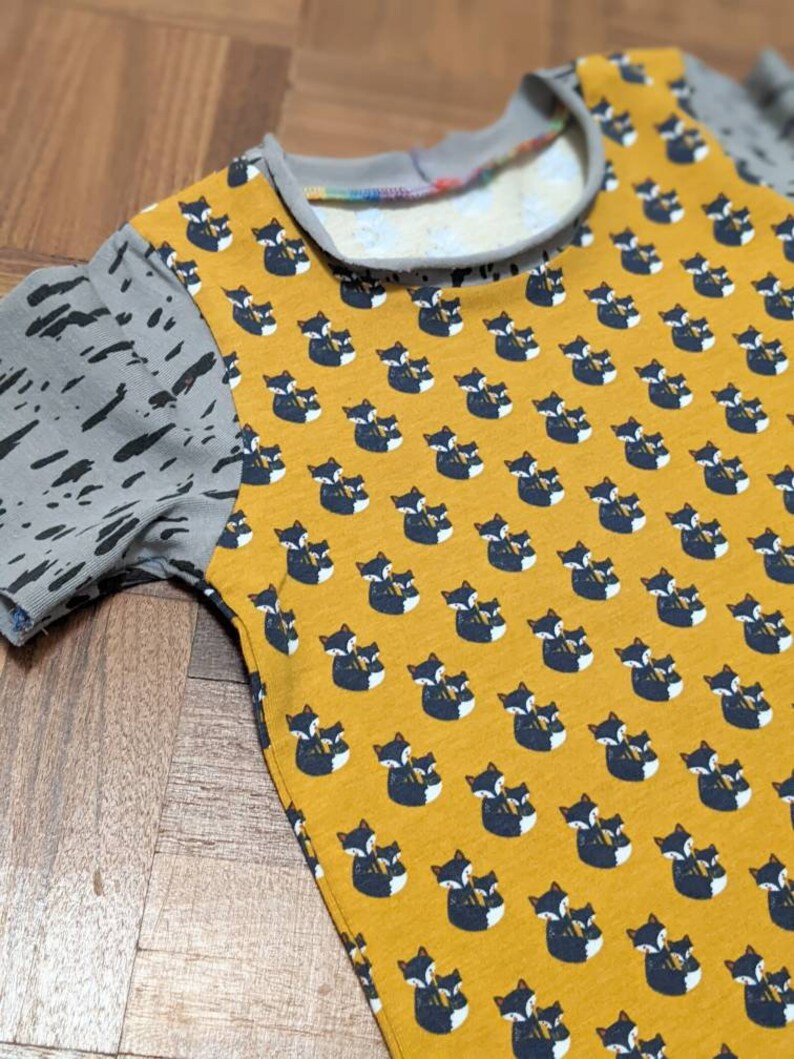 K&ouml;nnte beinhalten: Ein gelbes T-Shirt mit kurzen &Auml;rmeln und einem Muster aus schwarzen und wei&szlig;en F&uuml;chsen. Die &Auml;rmel sind aus grauem Stoff mit einem schwarzen Spritzmuster.