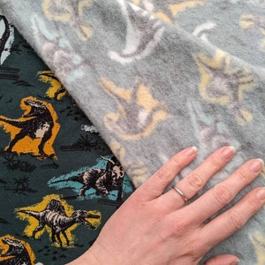 May include: A hand wearing a silver ring rests on a green fabric with a dinosaur pattern. The fabric is partially covered by a gray fabric with a white and yellow dinosaur pattern.