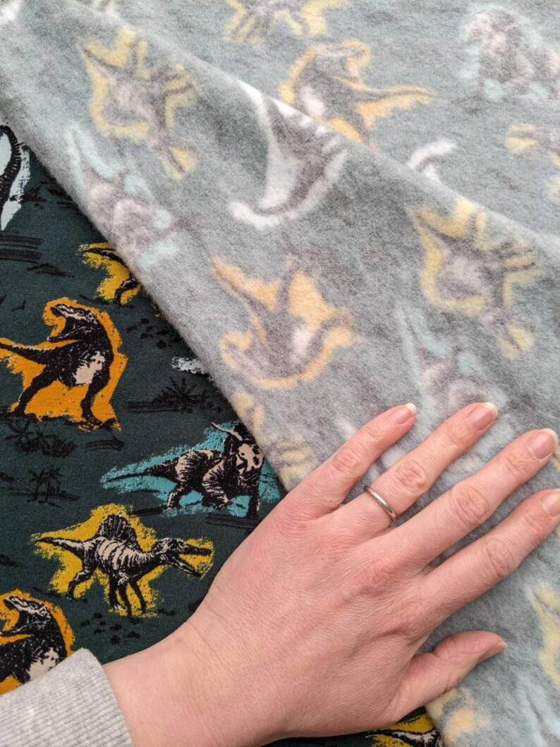 May include: A hand wearing a silver ring rests on a green fabric with a dinosaur pattern. The fabric is partially covered by a gray fabric with a white and yellow dinosaur pattern.