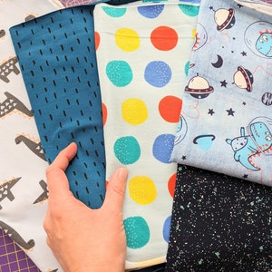 May include: A selection of colorful fabric swatches. Designs include dinosaurs, dotted patterns in various colors, and a space-themed print with dinosaurs in space suits. The fabrics are in shades of blue, white, and black.