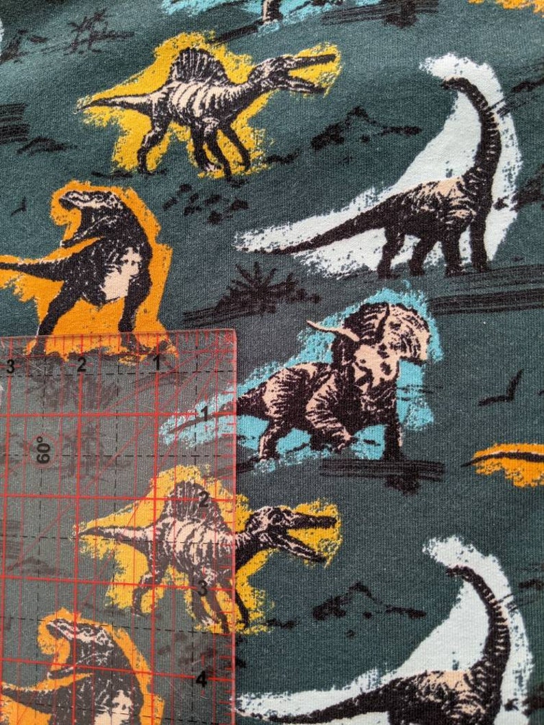 May include: A teal fabric with a repeating pattern of dinosaurs in various colors, including black, white, yellow, and blue. The dinosaurs are outlined in black and have a cartoon-like appearance.