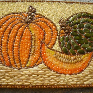 Könnte beinhalten: Ein Mosaikkunstwerk mit zwei Kürbissen, einem ganzen und einem geschnittenen, aus verschiedenen Samen und Körnern gefertigt. Die Kürbisse sind orange und grün, vor einem Hintergrund aus kleinen, beigen Samen. Ein einzigartiges, herbstliches Stück.