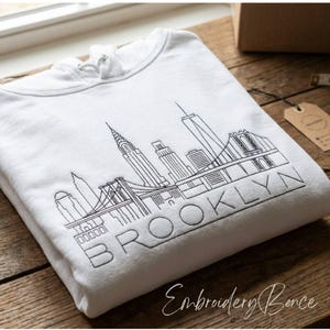 Puede incluir: Sudadera con capucha blanca con un contorno bordado en negro del horizonte de Brooklyn, incluyendo el puente de Brooklyn y la palabra "BROOKLYN". La sudadera está doblada sobre una superficie de madera.
