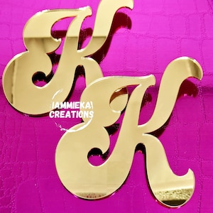 May include: Two gold-colored mirrored letter "K" cutouts. The letters are displayed on a textured, bright pink background. The text "IAMMIEKA! CREATIONS" is printed in white on the bottom left of the image.