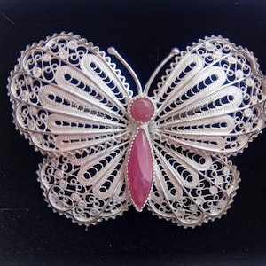 May include: A silver filigree butterfly brooch with a pink gemstone centre. The brooch has intricate openwork detailing on the wings and a delicate, lacy appearance. The butterfly's body is a teardrop-shaped pink gemstone.