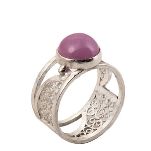 May include: A silver ring with a large, round, light purple gemstone. The ring has an openwork design with intricate patterns on the band. The gemstone is set in a simple bezel setting, and the ring is displayed against a white background.
