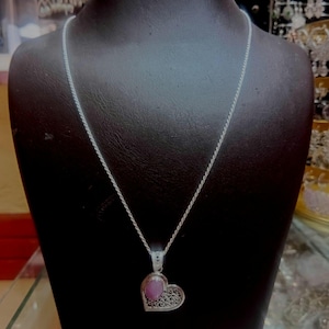 May include: A silver necklace with a heart-shaped pendant. The pendant has a pink gemstone set within a filigree heart design. The necklace is displayed on a black mannequin, highlighting the jewellery's details.