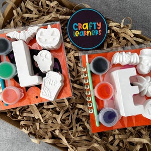 May include: Two craft kits in clear plastic packaging featuring white plaster shapes of letters L and E, ghosts, bats, pumpkins, a skull, and a tombstone. Each kit includes a paintbrush, small paint pots in various colours, and a Crafty Learners logo.