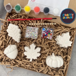May include: A craft kit with white ceramic shapes of leaves, an acorn, a pumpkin, and a maple leaf. The kit includes paint pots, glitter, and rhinestones. The box has the text "Crafty Learners" on it.