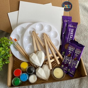 May include: A craft kit containing two white canvases, two wooden easels, a white paint palette, four paint pots, two white heart-shaped ceramic ornaments, a tea light candle, and two Cadbury chocolate bars.