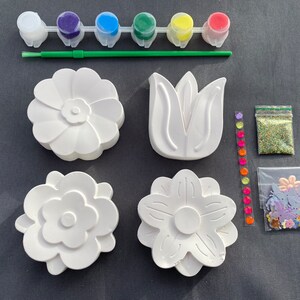 May include: A craft kit for painting ceramic flower shapes. The kit includes five small paint pots in red, green, blue, yellow, and purple, a paintbrush, a bag of glitter, and a bag of colorful flower-shaped embellishments.