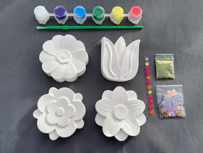 May include: A craft kit for painting ceramic flower shapes. The kit includes five small paint pots in red, green, blue, yellow, and purple, a paintbrush, a bag of glitter, and a bag of colorful flower-shaped embellishments.