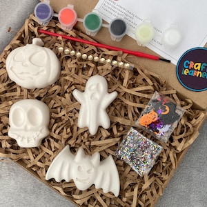 May include: A Halloween craft kit in a cardboard box containing four white ceramic figures: a pumpkin, a skull, a ghost, and a bat. The kit includes six small paint pots, a red paintbrush, a string of beads, and two bags of colorful confetti.