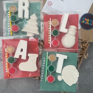May include: Christmas-themed craft kits with the letters R, A, L, and T, along with festive shapes like a tree, stocking, snowman, and Santa. Each kit includes paint pots, a brush, and decorative gems. The kits are packaged in clear plastic bags.