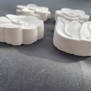 May include: Three white plaster flower-shaped ornaments. The ornaments are in the shape of a simple flower, a stylized flower, and a tulip.