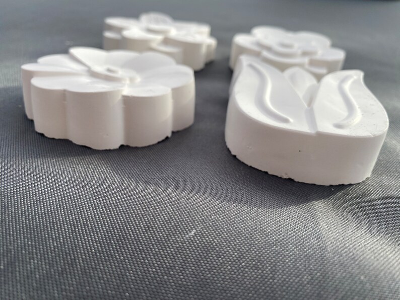 May include: Three white plaster flower-shaped ornaments. The ornaments are in the shape of a simple flower, a stylized flower, and a tulip.