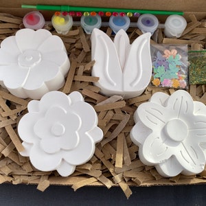 May include: A craft kit with three white ceramic flower shapes, a tulip shape, and a small bag of glitter. The kit also includes paint pots and a paintbrush.