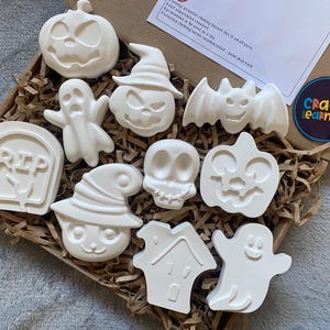 May include: A set of ten white plaster Halloween-themed figures including pumpkins, ghosts, a skull, a bat, a tombstone with the text RIP, and a house. These unpainted shapes are arranged in a cardboard box filled with brown paper filler strips.