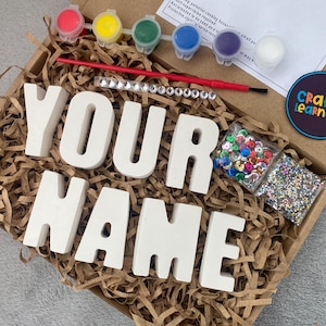 May include: A craft kit with white ceramic letters spelling "YOUR NAME" in a brown paper shred box. The kit includes paint pots in red, yellow, blue, green, and purple, a paintbrush, a bag of colorful sequins, and a bag of glitter.