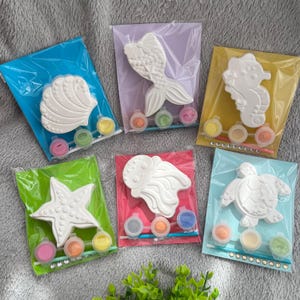 May include: Five paint-your-own kits featuring sea creatures: a seashell, mermaid tail, seahorse, starfish, and turtle. Each kit includes a white plaster figure, paint pots, and a paintbrush, all packaged in colourful plastic bags.