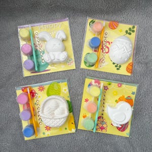May include: Four Easter-themed craft kits. Each kit includes a white plaster figure, paint pots in various colors, and a paintbrush. The figures include a bunny, an egg, a basket of eggs, and a chick. The packaging has the word "Happy".