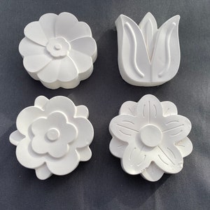 May include: Four white plaster flower shapes. Two of the shapes are simple flowers with five petals. One shape is a flower with seven petals. The last shape is a stylized tulip.