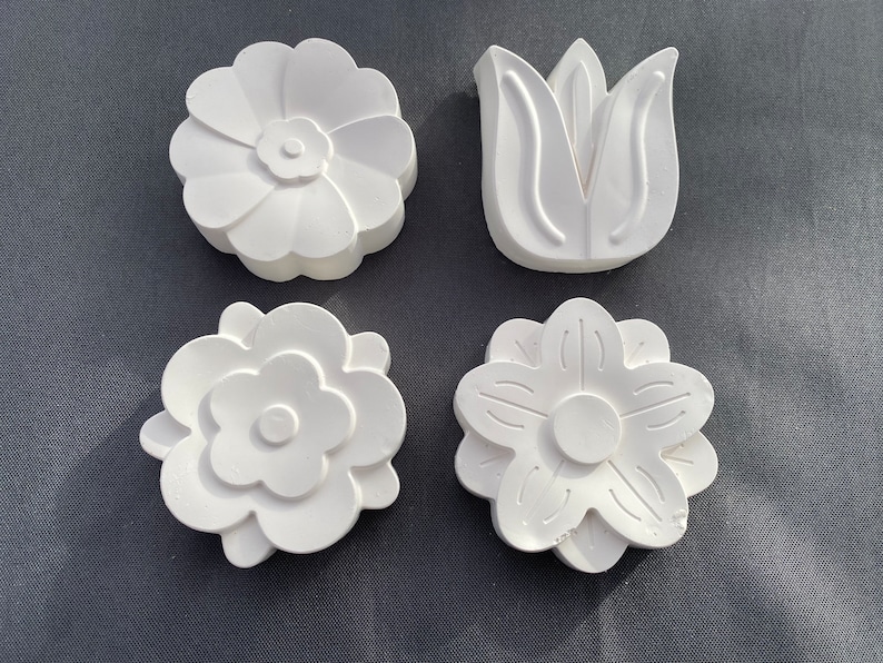 May include: Four white plaster flower shapes. Two of the shapes are simple flowers with five petals. One shape is a flower with seven petals. The last shape is a stylized tulip.