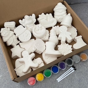 May include: A selection of unpainted ceramic Christmas ornaments in a cardboard box, featuring festive shapes like snowmen and gingerbread men. The set includes pots of paint in various colours and three paintbrushes, ideal for a creative holiday project.