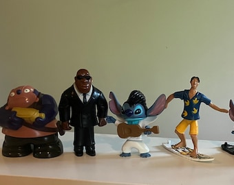 May include: A set of seven plastic figurines, including a brown and black figure, a black figure wearing a suit, a blue and white figure holding a guitar, a blue figure on a surfboard, a blue figure on a surfboard wearing a red suit, a blue figure on a surfboard wearing a brown and white outfit, and a blue figure on a surfboard wearing a blue and white outfit.