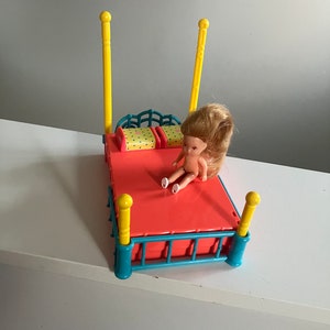 May include: A colourful vintage doll's bed. The bed frame is coral with teal accents and yellow posts. Two small pillows with yellow polka dots are on the bed. A small doll with blonde hair is sitting on the bed.
