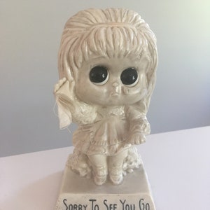 May include: A white ceramic figurine of a girl with large eyes and a sad expression. She is holding a handkerchief to her face and is sitting on a base that reads "Sorry To See You Go".