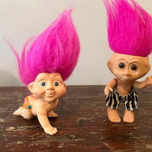 May include: Two pink-haired troll dolls. One is a baby troll wearing a nappy and crawling. The other is a standing troll wearing black and white striped shorts.