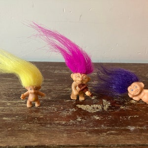 May include: Three small troll dolls with colourful hair. The troll on the left has yellow hair, the middle troll has pink hair and is playing a violin, and the troll on the right has purple hair and is holding a yellow flower.