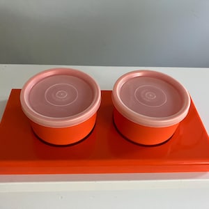May include: Two orange containers with pink lids sit on an orange tray. The containers are round and have a slight lip around the top.