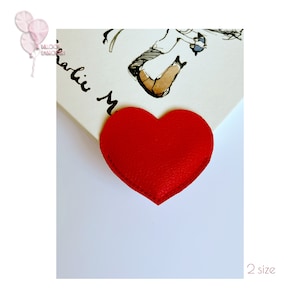 May include: A red heart-shaped leather bookmark with a stitched edge. The bookmark is on a white surface with the text "Charlie Mack" written on it.