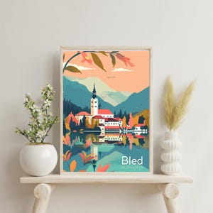 Op de afbeelding: Een ingelijste kunstafdruk met een kleurrijke illustratie van Bled, Slovenië. Het kunstwerk toont een kerk met een hoge toren aan een meer, omringd door bergen en bomen. De tekst "Bled Slovenië" staat onderaan.