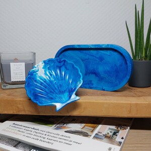 Op de afbeelding: Twee blauwe en witte hars trays, een in de vorm van een schelp en de andere ovaal. De trays staan op een houten oppervlak met een kaars en een potplant op de achtergrond.