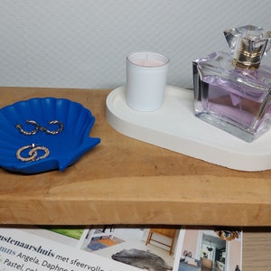 Op de afbeelding: Een wit ovaal dienblad met een witte kaars en een fles parfum. Het dienblad ligt op een houten oppervlak met een blauwe schelpvormige schaal met gouden oorbellen.