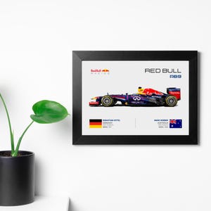 May include: A framed print of a Red Bull Racing Formula One car, the RB9, with the drivers Sebastian Vettel and Mark Webber. The car is blue, purple, and red with the Red Bull logo and sponsors' logos. The print includes the drivers' names, nationalities, and race results.