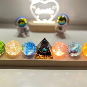 Pode incluir: Sete esferas de vidro, cada uma com uma cor e brilho diferentes, são dispostas em uma base de madeira. Uma pirâmide dourada com um design preto e verde fica no centro da base.